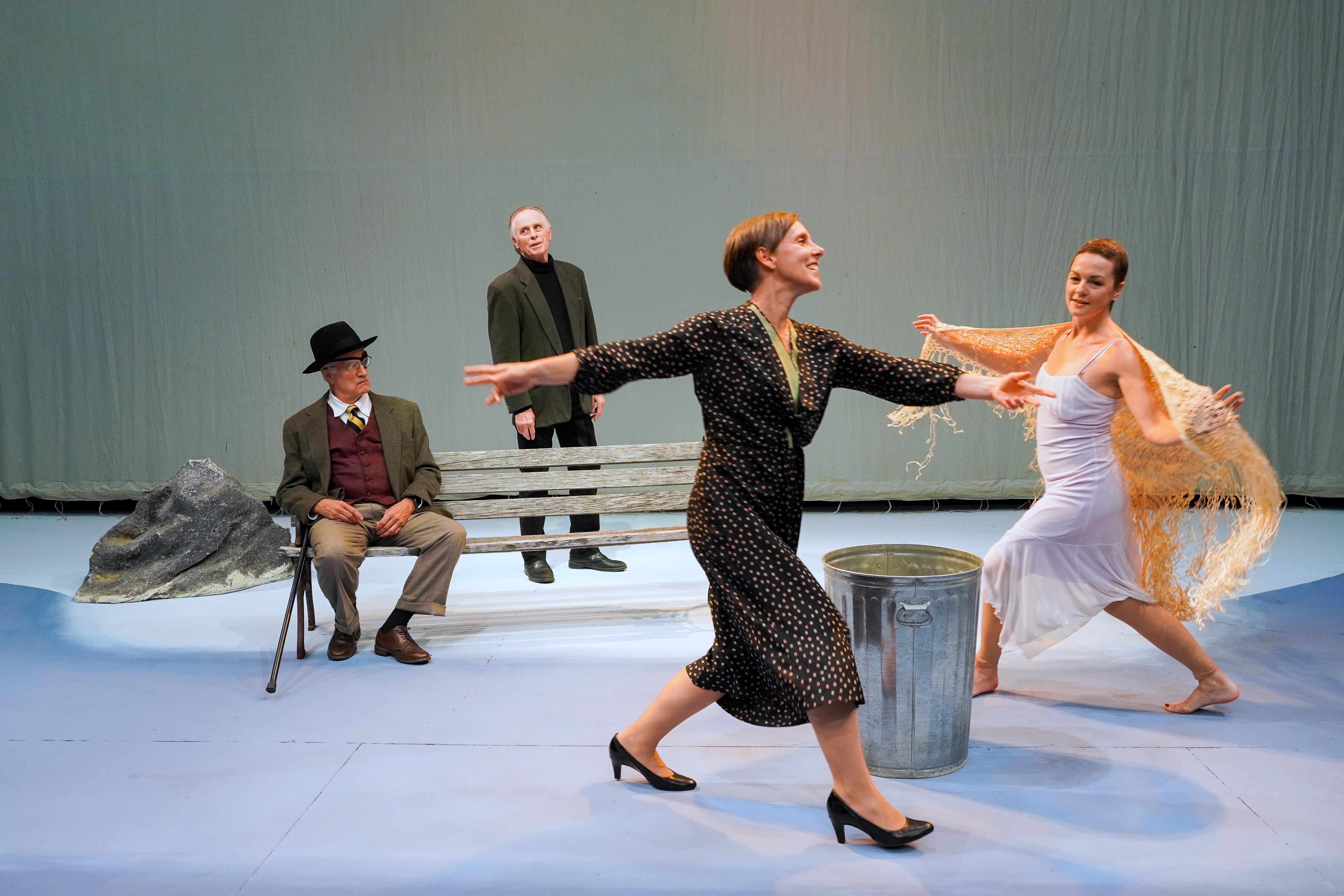 pale blue sky drop behind actors, large rock on left, sitting on bench in suit & vest, eye patch & glasses, black hat is James Joyce, standing behind him in black slacks, turtleneck & jacket is Samuel Beckett, in foreground on right dancing while circling a trashcan are actors playing Suzanne & Lucia, in dresses, in a skipping pose